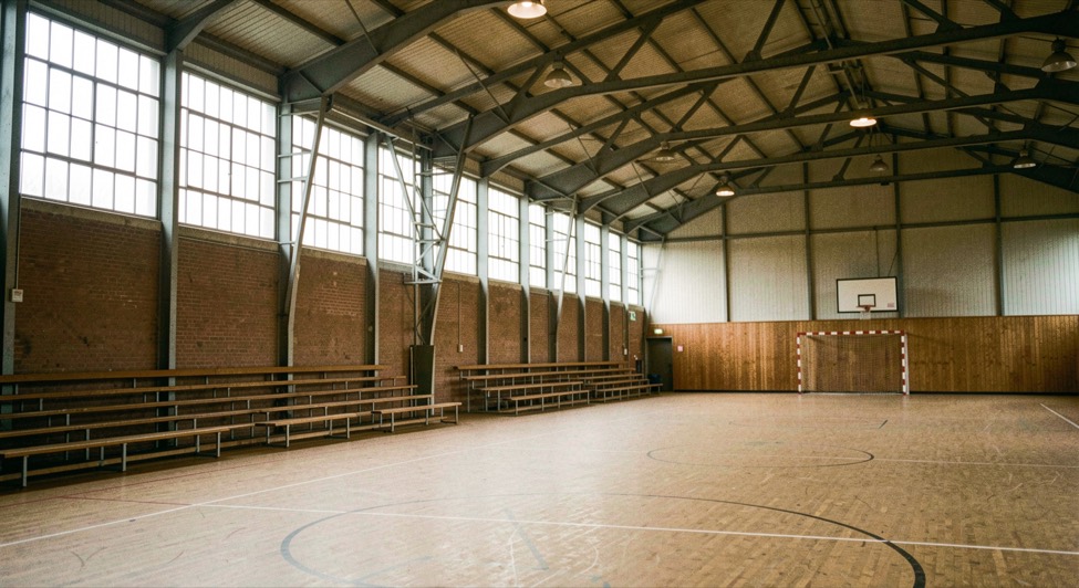 Het interieur van een grote naoorlogse Nederlandse sporthal met een handbalveld, hoge ramen en lege houten banken langs de wand